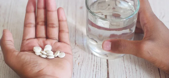 Hands holding white tablets and a glass of water over a wooden table