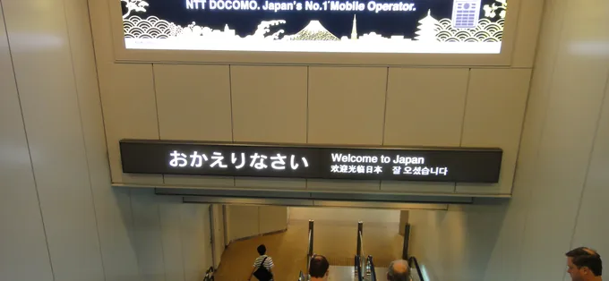 The welcome board at the arrival gate of Narita Airport, with travellers going down the escalator