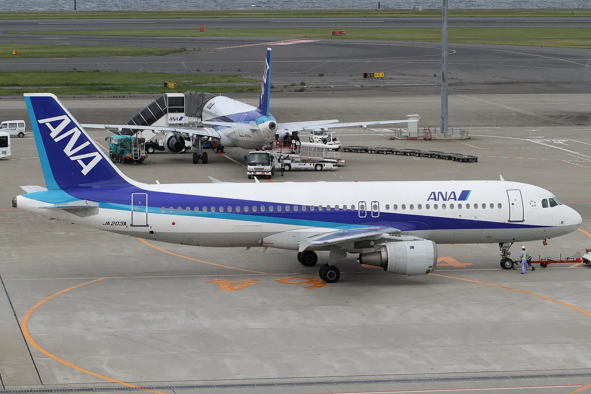 ANA Airbus A320 aircraft parked at a terminal gate with ground support vehicles and another ANA aircraft in the background