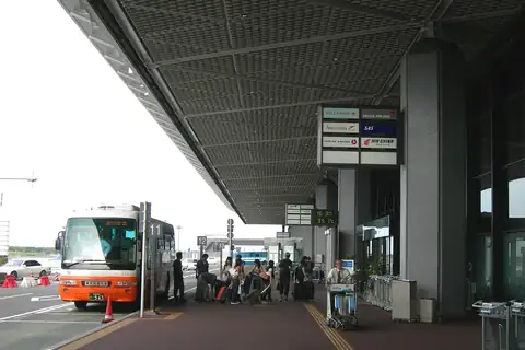 An orange and white airport limousine bus parked under a terminal canopy with passengers and luggage, Narita Airport