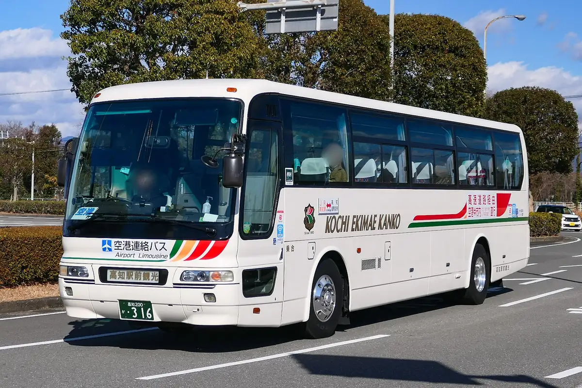 生け垣と木々沿いの道路を走る、白い高知駅前観光の空港リムジンバス