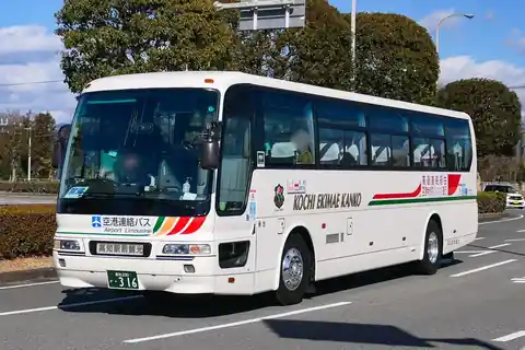 A white Kochi Ekimae Kanko airport limousine bus on a road beside hedges and trees
