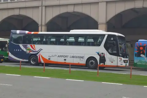 A Kyusanko airport limousine bus with a bear mascot on its side, beside an arched overpass