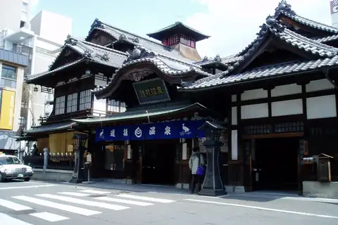 Dogo Onsen Honkan, a tiered wooden bathhouse with dark tiled roofs and a blue entrance curtain in Matsuyama