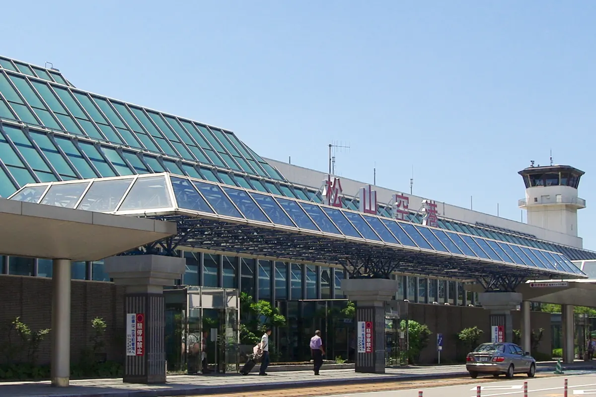 Exterior of Matsuyama Airport terminal building with a glass facade, control tower, and a sign reading Matsuyama Airport