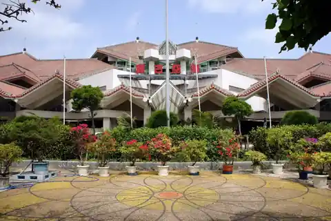 Red-tiled terminal building of Miyako Airport with flagpoles, flowering shrubs, and potted plants in front