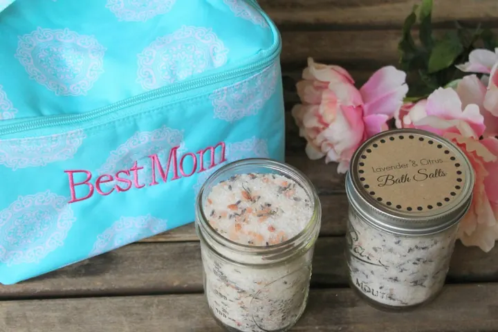 Two glass jars of bath salts labeled Lavender and Citrus beside pink flowers and a turquoise bag embroidered Best Mom