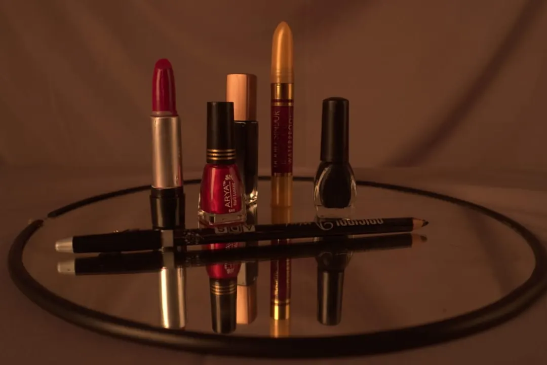 A red lipstick, two nail polish bottles, a yellow cosmetic stick, and an eyeliner pencil on a round mirror
