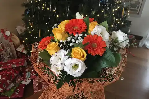 A bouquet of orange, yellow, and white flowers wrapped in orange mesh paper, with a Christmas tree in the background