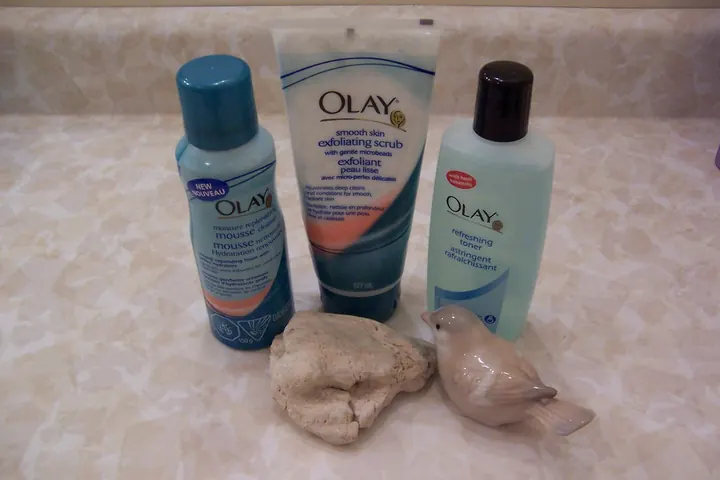 Olay mousse cleanser, exfoliating scrub tube and toner bottle on a counter, with a small bird figurine