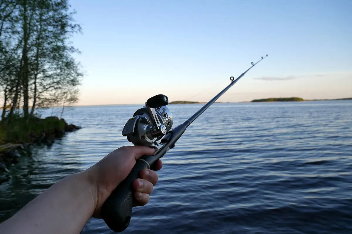 A hand holding a fishing rod with a spinning reel, with trees and water in the background