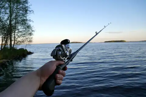 A hand holding a fishing rod with a spinning reel, with trees and water in the background