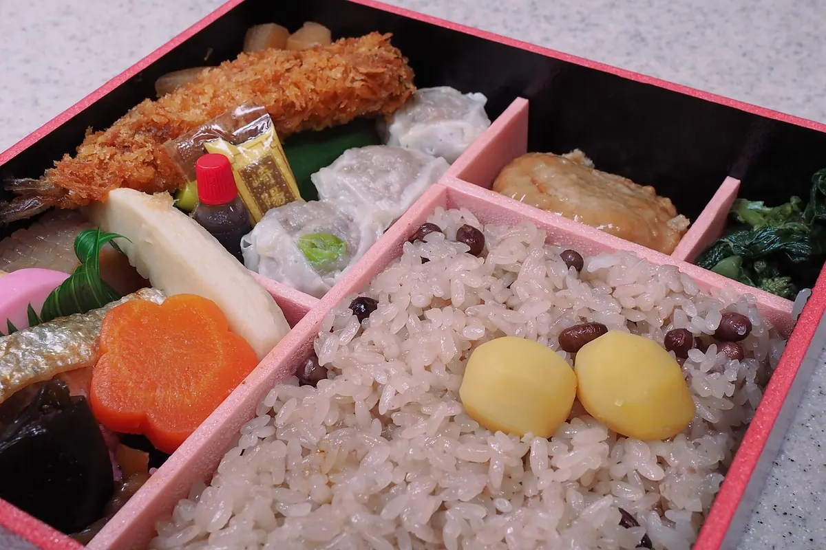 A Japanese bento box with rice, black beans, chestnuts, fried shrimp, grilled fish, and vegetables in compartments