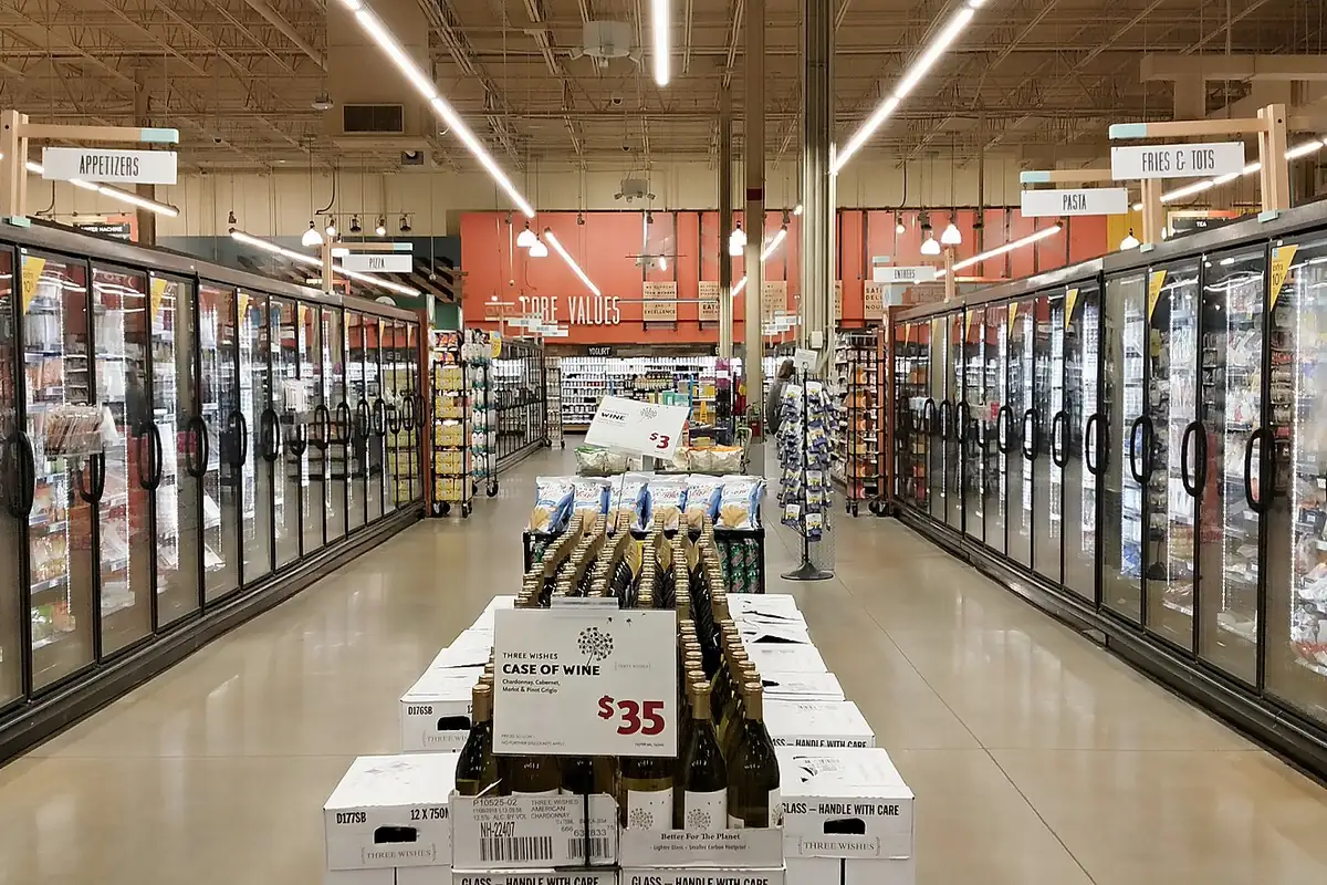 Rows of glass-door freezers stocked with frozen food in a supermarket aisle, with a wine display in the center
