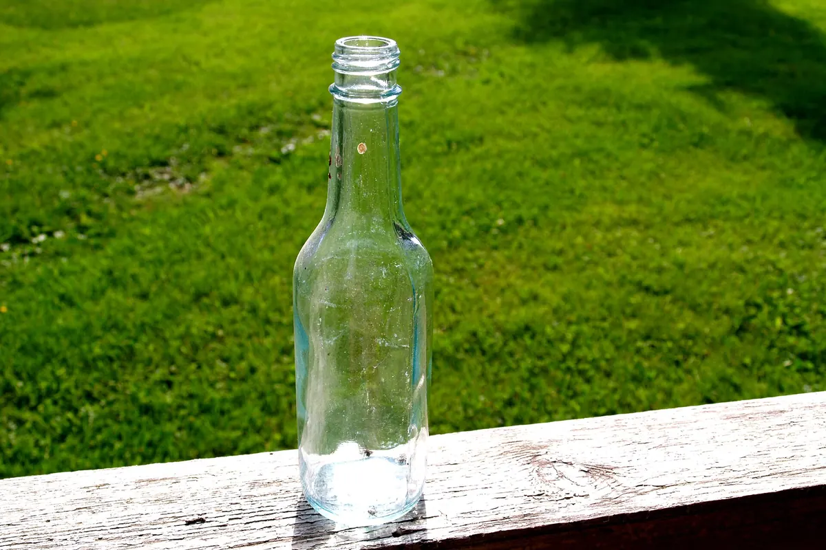 A clear empty glass bottle standing on a wooden rail with grass behind it