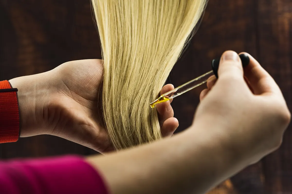 金色のヘアオイルをスポイト式のアプリケーターで金髪の毛先に塗る手
