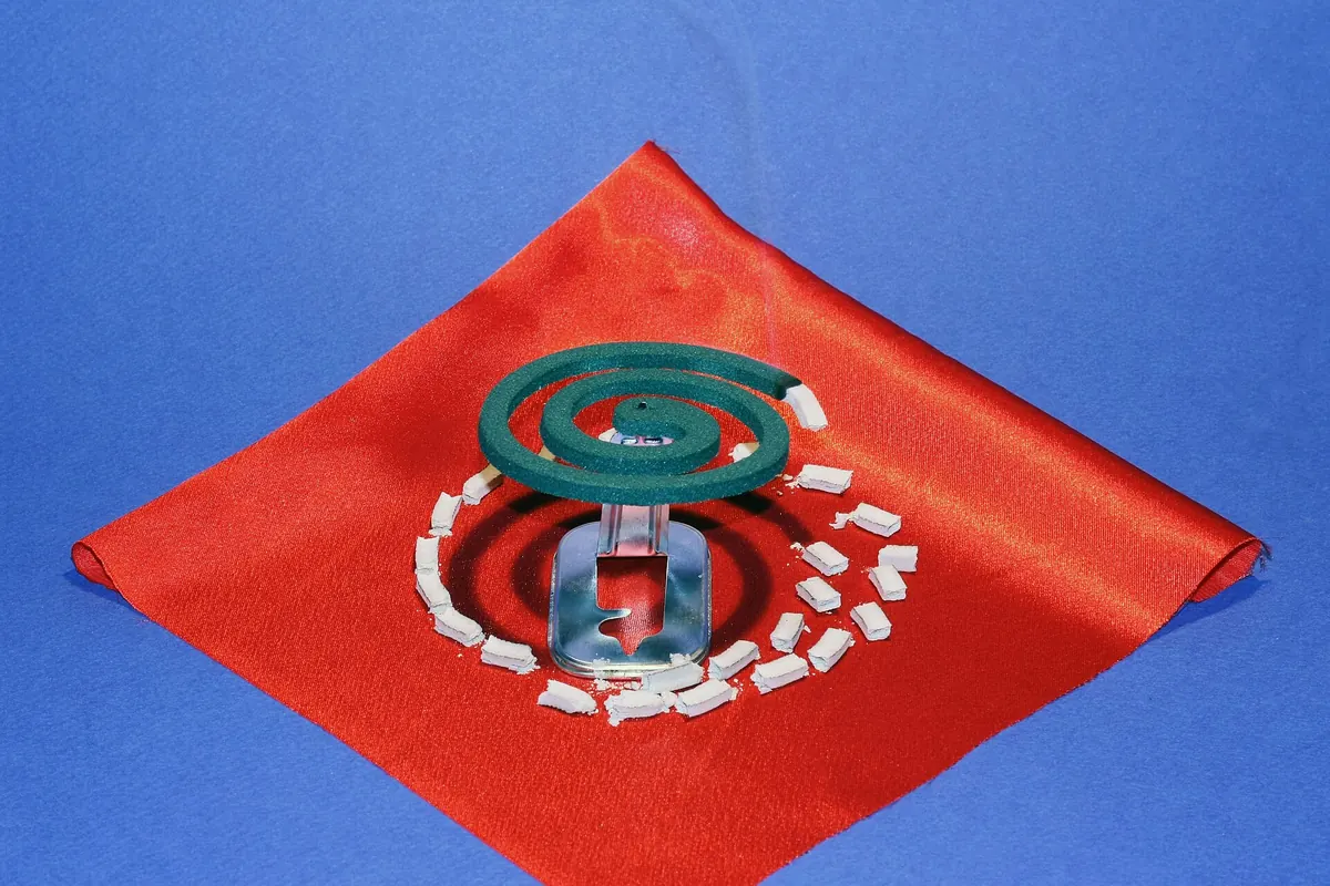 A green coiled mosquito repellent incense on a metal stand with scattered ash on a red cloth over a blue background