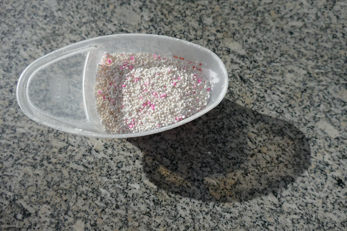 Laundry detergent granules with pink specks in a white measuring cup on a grey surface