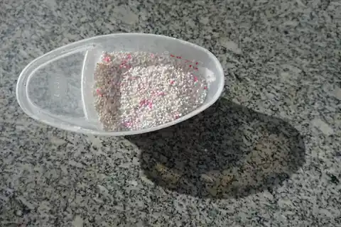Laundry detergent granules with pink specks in a white measuring cup on a grey surface