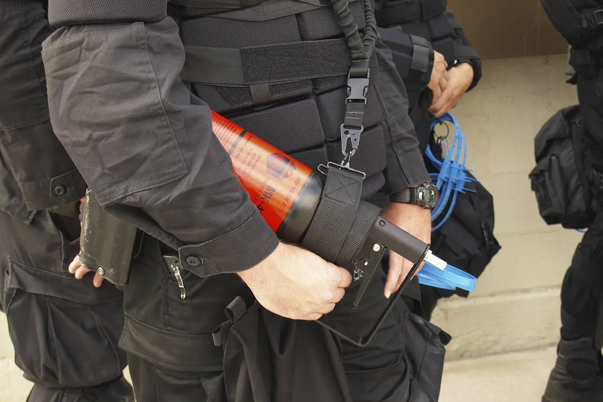 Security personnel in black tactical gear holding a red-orange pepper spray canister at the waist