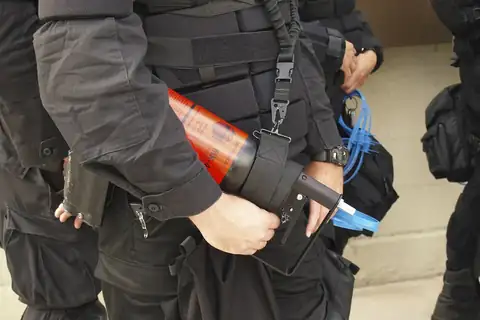 Security personnel in black tactical gear holding a red-orange pepper spray canister at the waist