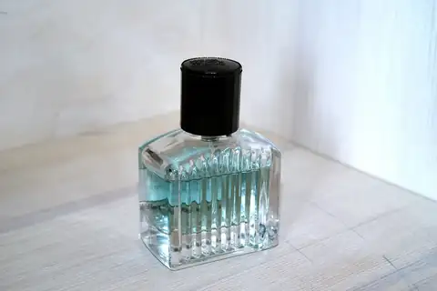 A square ribbed glass perfume bottle with a black cap and pale blue liquid on a wooden surface