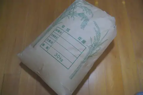 A beige rice sack with a green rice plant design and Japanese text, on a wooden floor