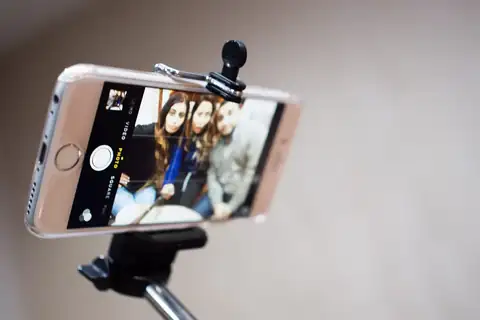 A smartphone clamped to a selfie stick, with a camera app showing three people on screen