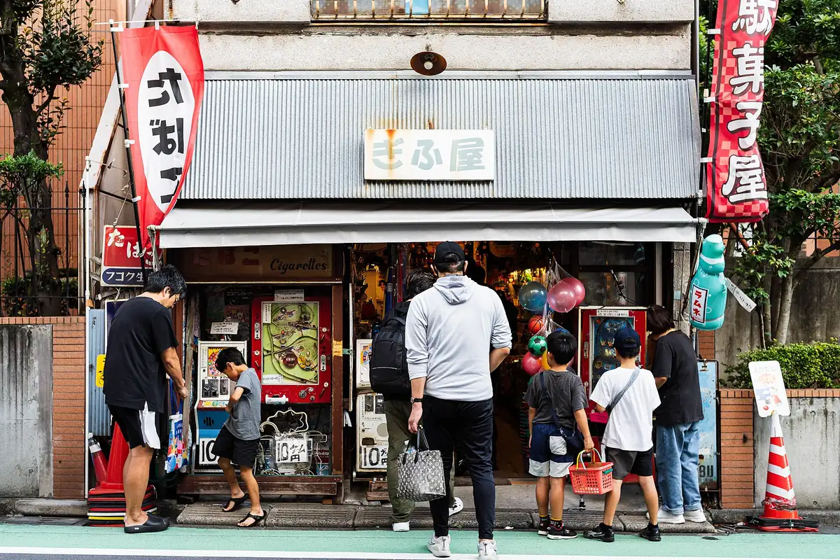 Storefront of a Japanese candy shop with red banners, balloons at the entrance, and people standing outside
