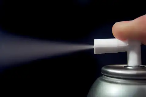 Finger pressing the nozzle of a silver aerosol can, with a fine mist against a dark background