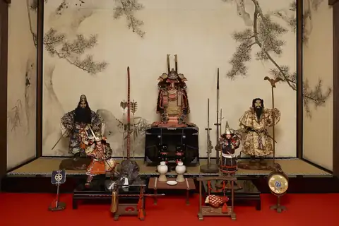 Armored samurai dolls with helmets, bows, spears, and swords in front of a painted pine screen