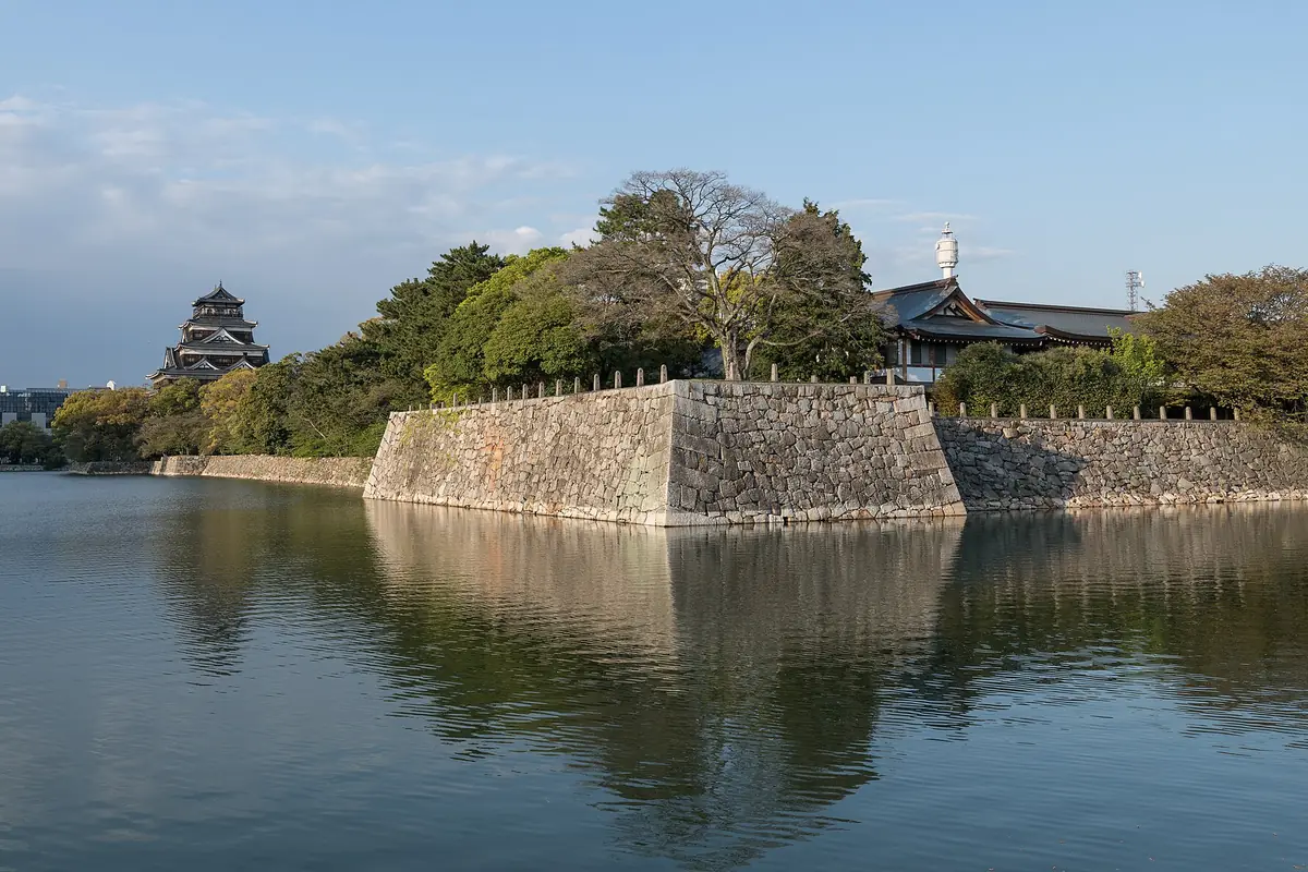 Stone wall along the moat of Hiroshima Castle, with the dark-roofed keep tower behind the trees on the left