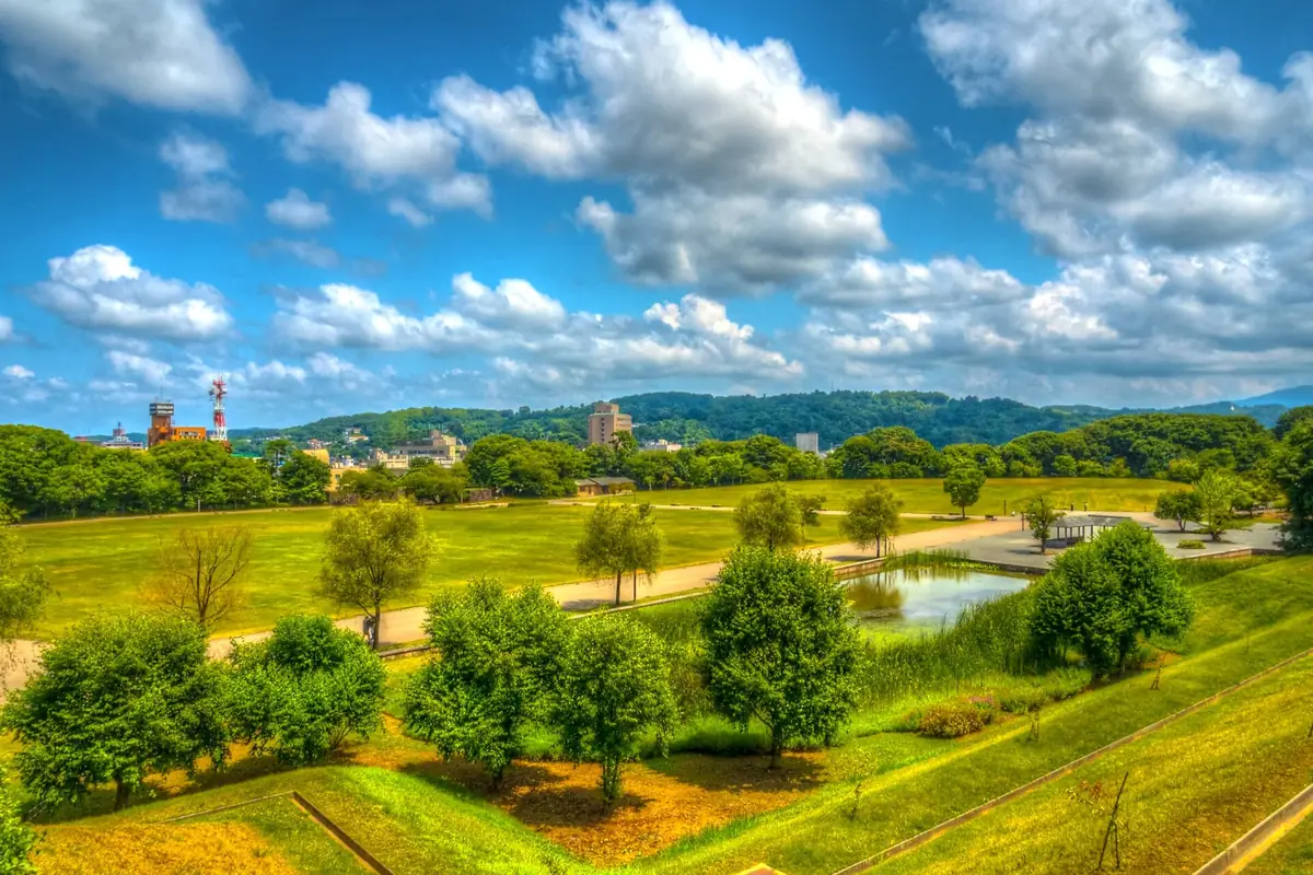 Lush green lawn, trees and a small pond in Kanazawa Castle Park, with wooded hills in the distance