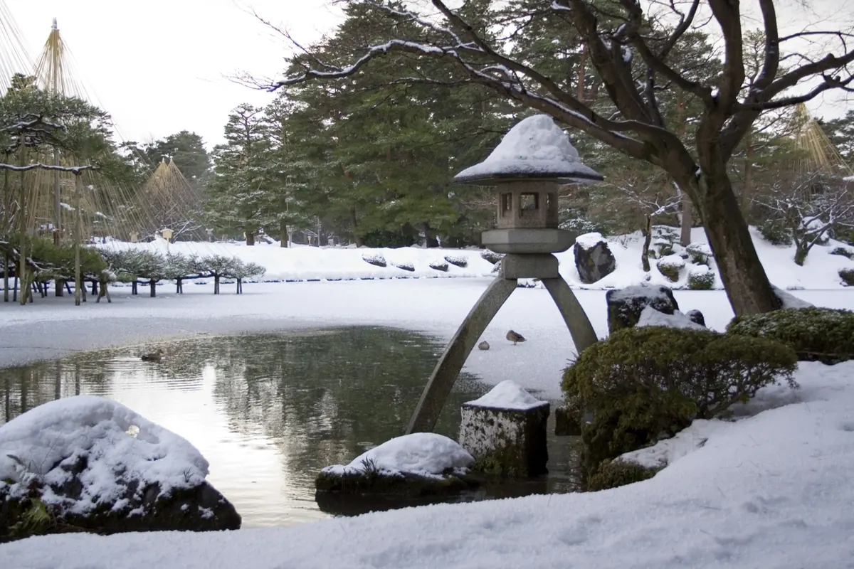 Snow-covered stone lantern beside a pond in Kenroku-en Garden, with yukitsuri ropes in the background