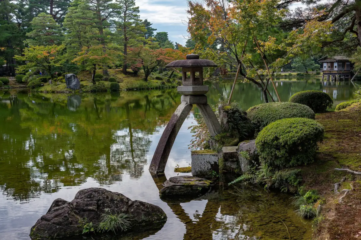 A stone lantern in a pond beside trimmed shrubs and trees with orange and yellow leaves at Kenroku-en Garden, Kanazawa
