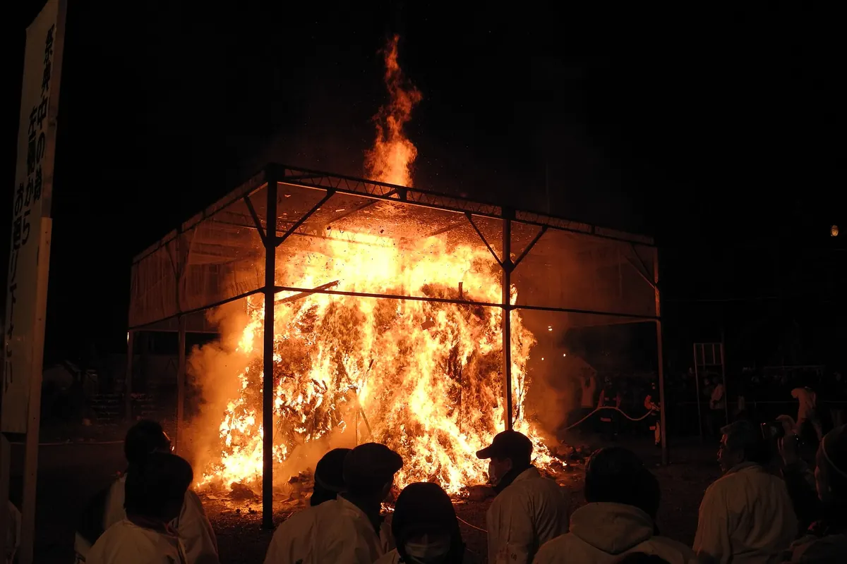 Large fire blazing inside a metal frame at night during a fire festival, with people in white watching in front