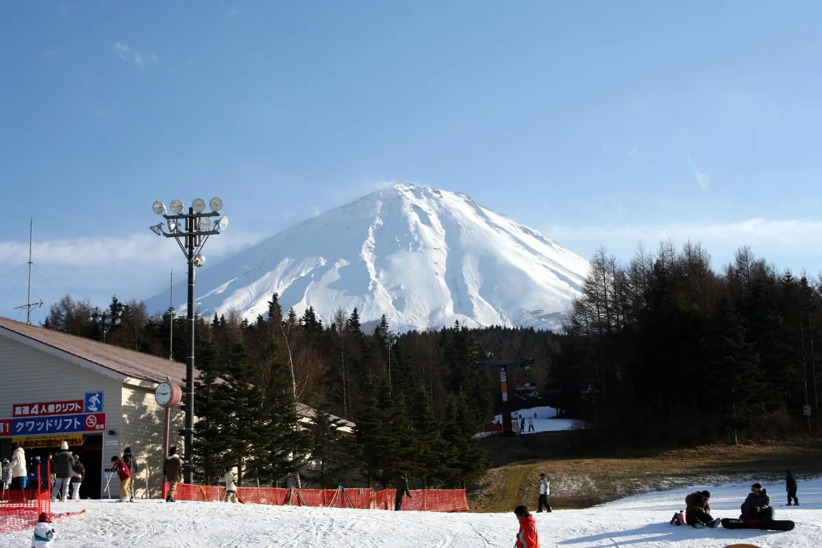 Snow-covered Mount Fuji above a forest, with a ski slope, a lift station and people on the snow in the foreground