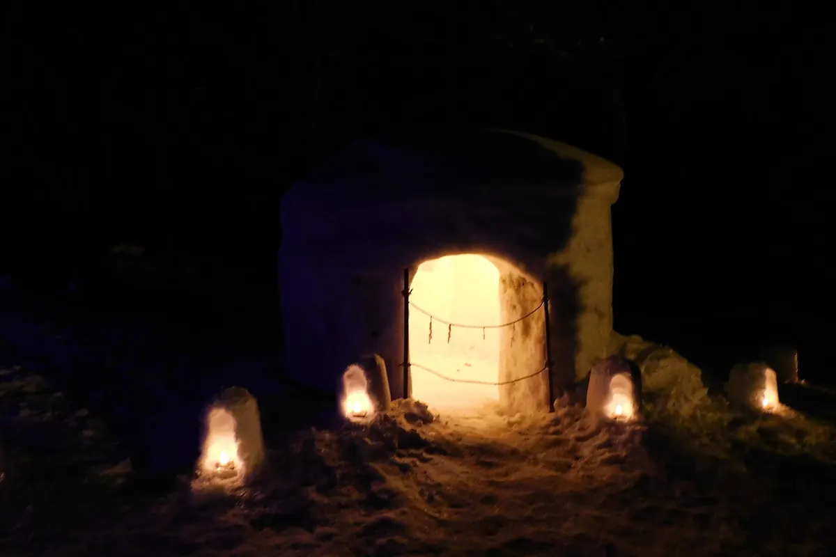 Snow hut (kamakura) glowing from inside at night, with small lit snow lanterns on the ground in front of it