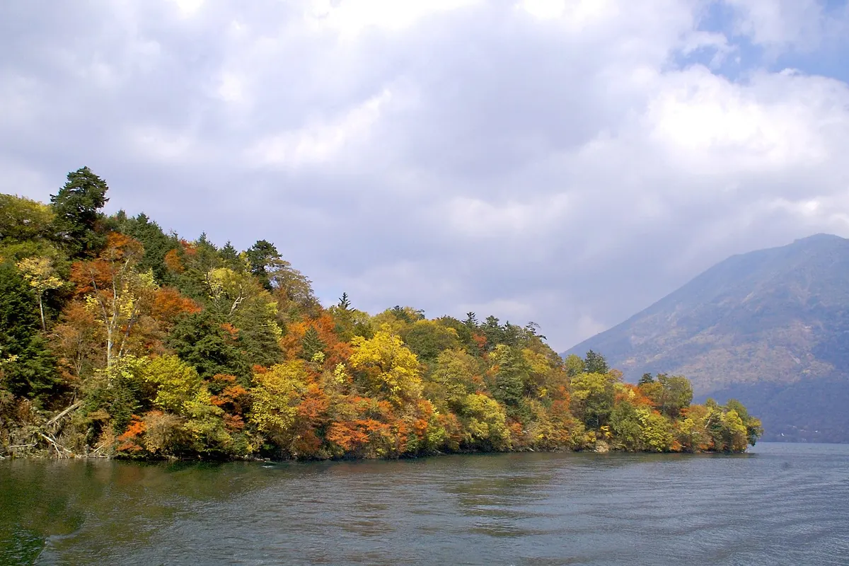 Autumn-colored trees along the shore of Lake Chuzenji, with a mountain rising on the right