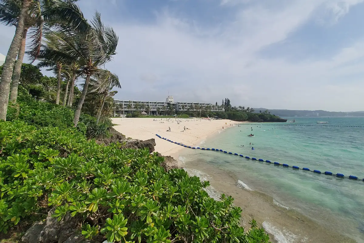 Moon Beach in Onna, Okinawa, seen past palm trees, with a hotel building, white sand, and a line of swimming buoys