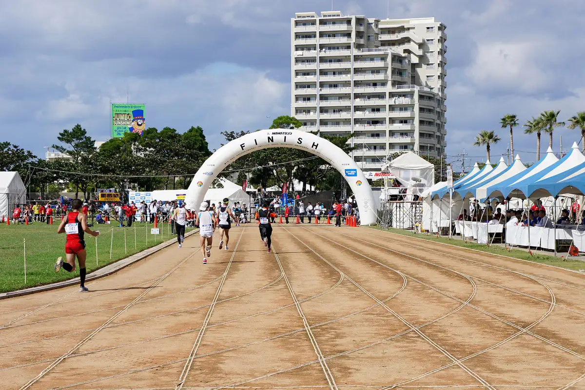 Runners on a track at the Naha Marathon, with a white arch and high-rise buildings behind