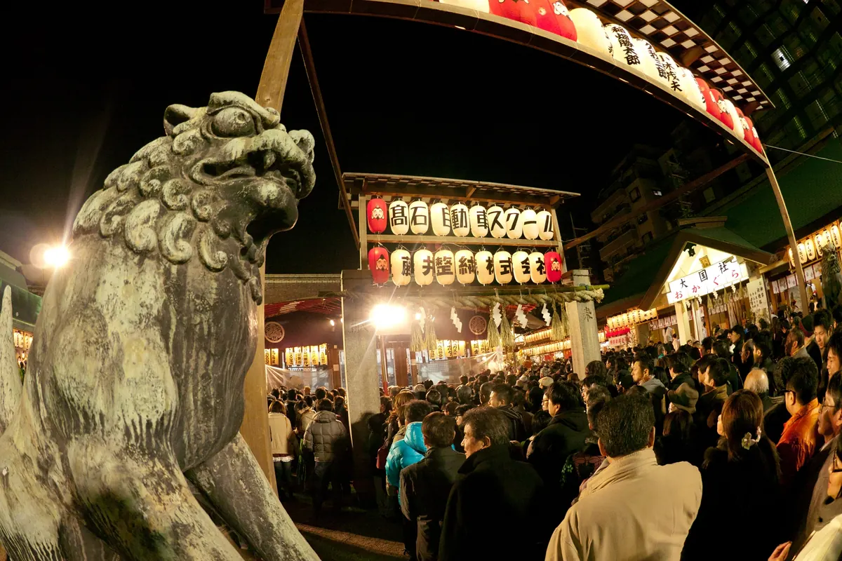 A stone guardian lion-dog statue, with a crowd and lantern-lit gateway behind it at night during Imamiya Ebisu Festival
