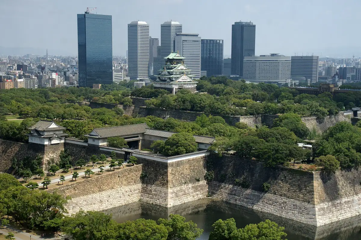 Osaka Castle's main tower rising above dense green trees and stone walls, with skyscrapers behind it