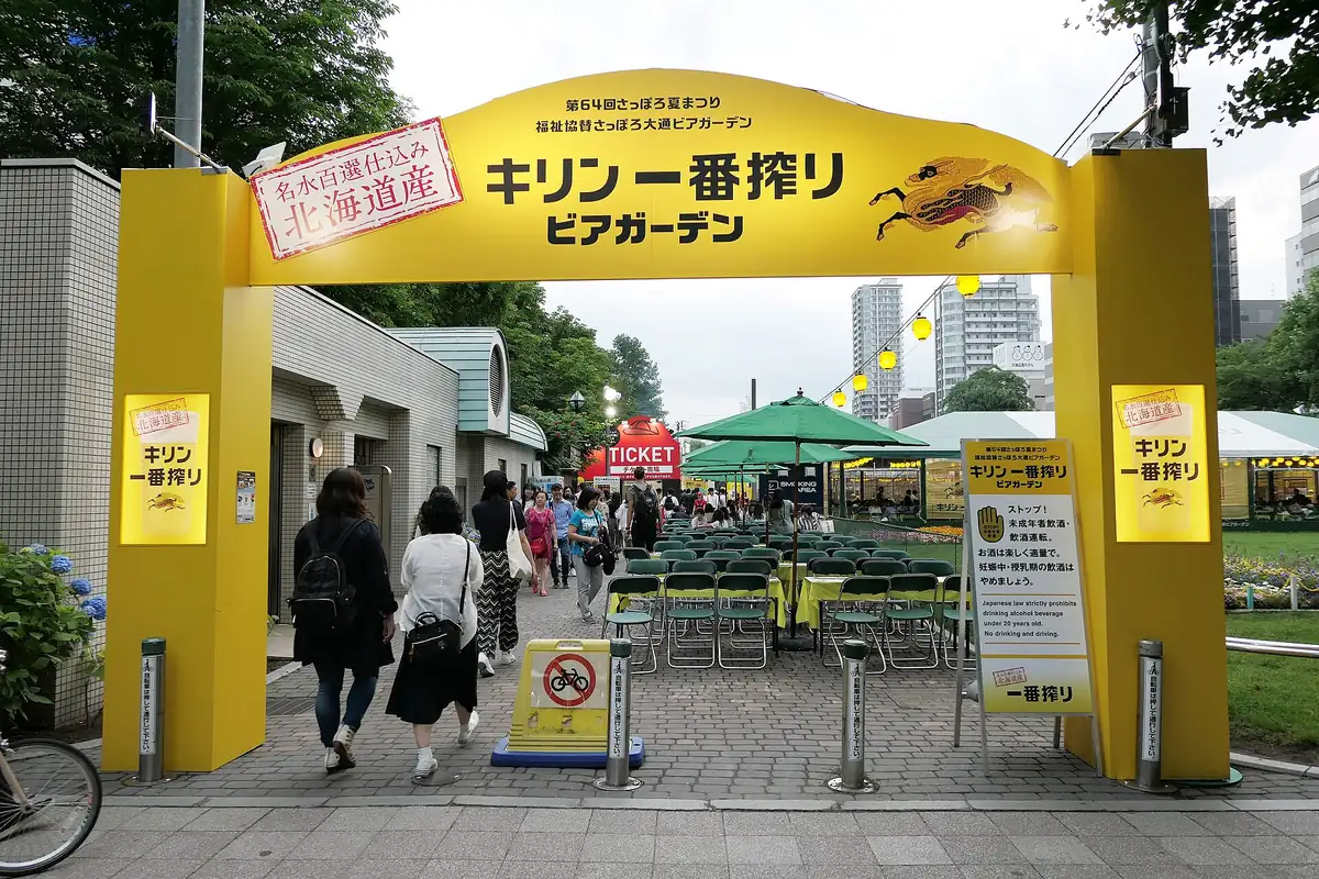 People walking under a yellow beer garden entrance arch with green umbrellas and tables behind it