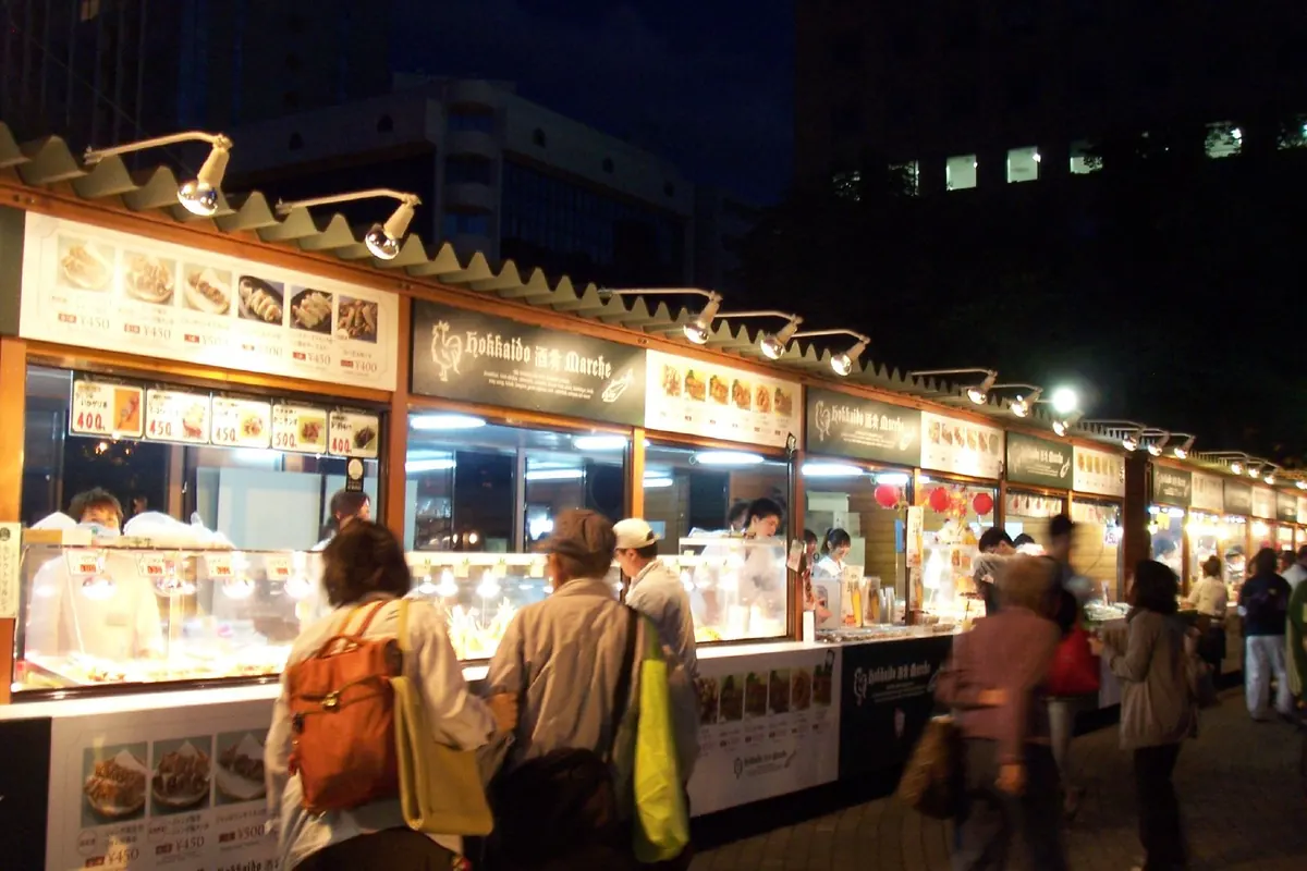 Lit food stalls at night with price menu boards at Sapporo Autumn Fest, with people standing and walking in front