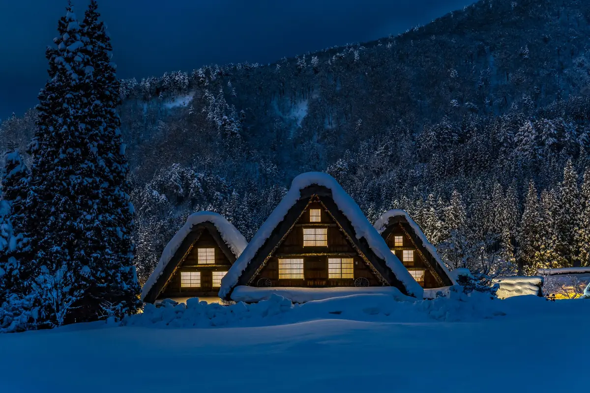 Three steep-roofed gassho-zukuri houses with snow on their roofs and lit windows at night