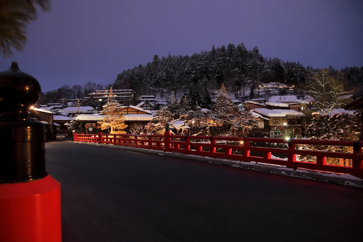 Red Nakabashi Bridge lit up at night in Takayama, with snow on the rooftops behind it