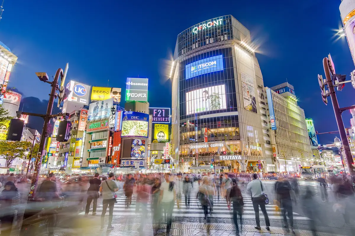 Blurred pedestrians crossing Shibuya Scramble Crossing, with the lit-up Q-Front building rising above them in Tokyo