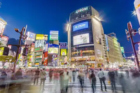 시부야 스크램블 교차로를 건너는 흐릿한 보행자들과, 그 위로 솟은 조명이 켜진 큐프론트 빌딩