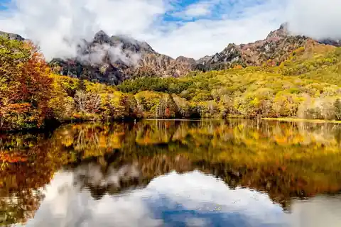 Mountains and clouds reflected in a calm pond, surrounded by red, orange, and yellow autumn foliage at Togakushi